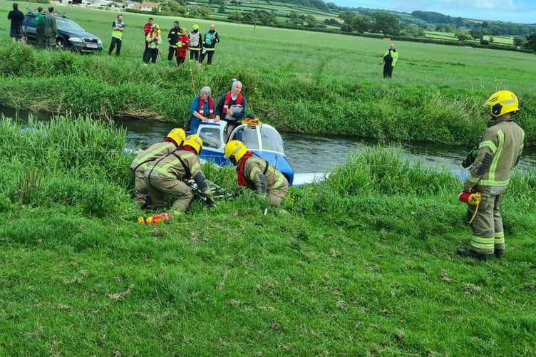 Pilot and passenger stand on a wing of their crashed aircraft in the River Axe while firefighters work on their rescue.